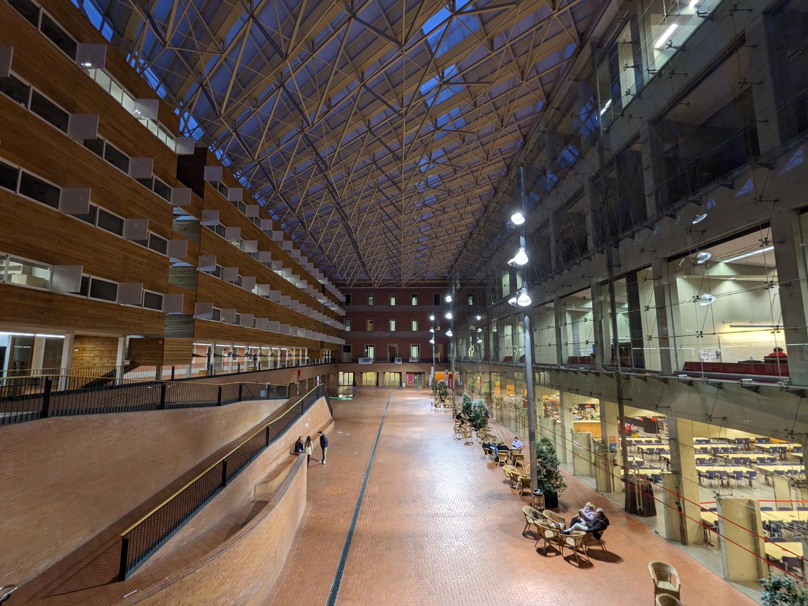 Two buildings with a inner floor that has a large roof. The left building has a wooden design, the right one has a large glass front in which you can see some lecture halls.